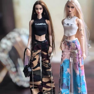 May include: Two fashion dolls wearing crop tops with "Calvin Klein" text. One doll has long brown hair and camouflage pants, the other has blonde hair and tie-dye pants. Both dolls wear sneakers and have accessories.