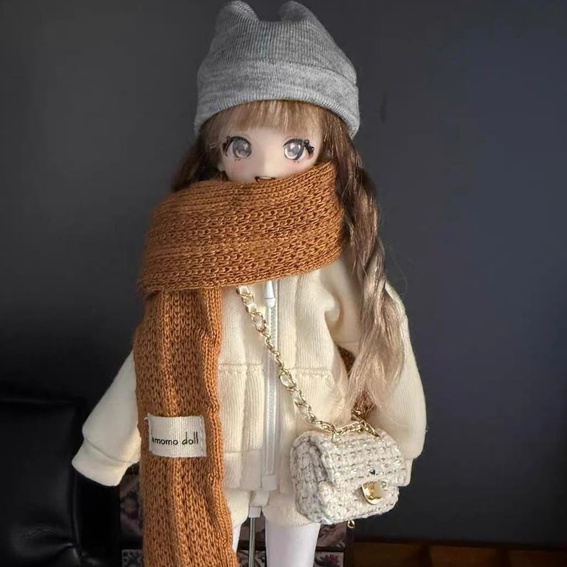 Puede incluir: Una mu&ntilde;eca con gorro gris, bufanda marr&oacute;n y chaqueta color crema. La mu&ntilde;eca tiene pelo largo casta&ntilde;o y lleva un bolso blanco peque&ntilde;o con cadena dorada. La bufanda tiene una etiqueta que dice "momo doll".