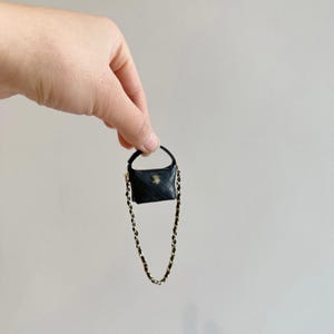 May include: A miniature black handbag with a quilted pattern and a gold-colored chain strap. The small purse has a curved handle and a gold-colored emblem on the front. The bag is held up by a hand.
