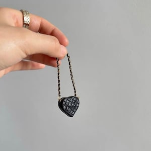 May include: A small, black heart-shaped purse with a quilted pattern. The purse has a gold chain strap and a small clasp. The purse is held up by a hand against a gray background. The purse is a miniature version of a classic handbag.