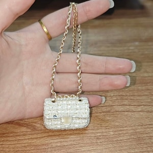Puede incluir: Un minibolso de tweed blanco con una cadena y un cierre dorados. El peque&ntilde;o bolso se sostiene en una mano, mostrando su delicado dise&ntilde;o y artesan&iacute;a. La textura y los detalles del bolso son claramente visibles.