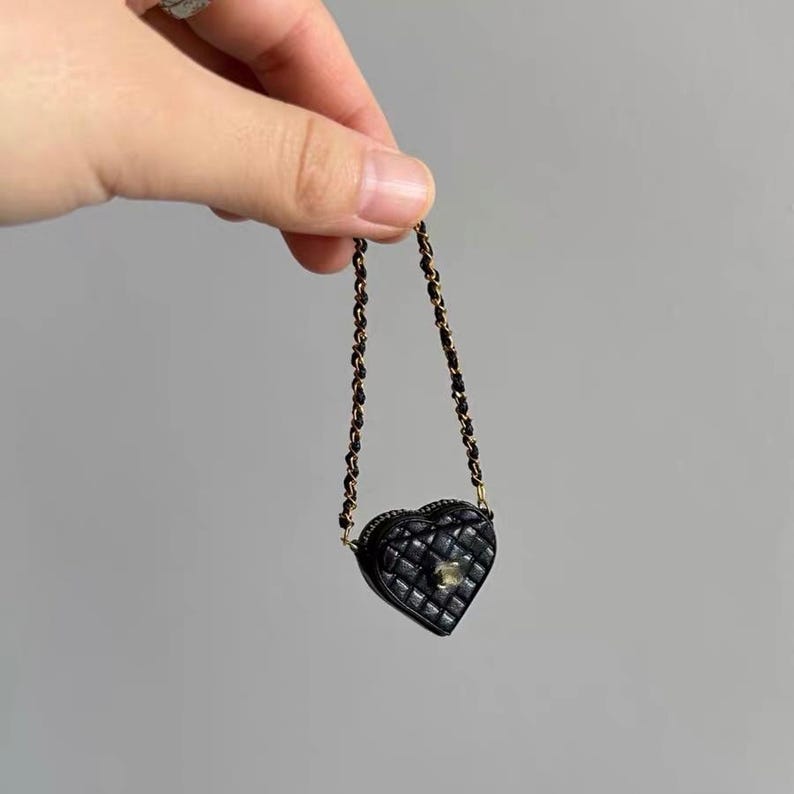 May include: A small, black heart-shaped purse with a quilted design. The purse has a gold chain strap and a gold clasp. The purse is held up by a hand against a gray background.