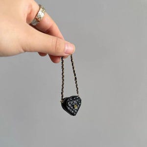 May include: A miniature black heart-shaped purse with a quilted pattern and a gold-colored clasp. The purse has a delicate chain strap. The purse is held up by a hand.