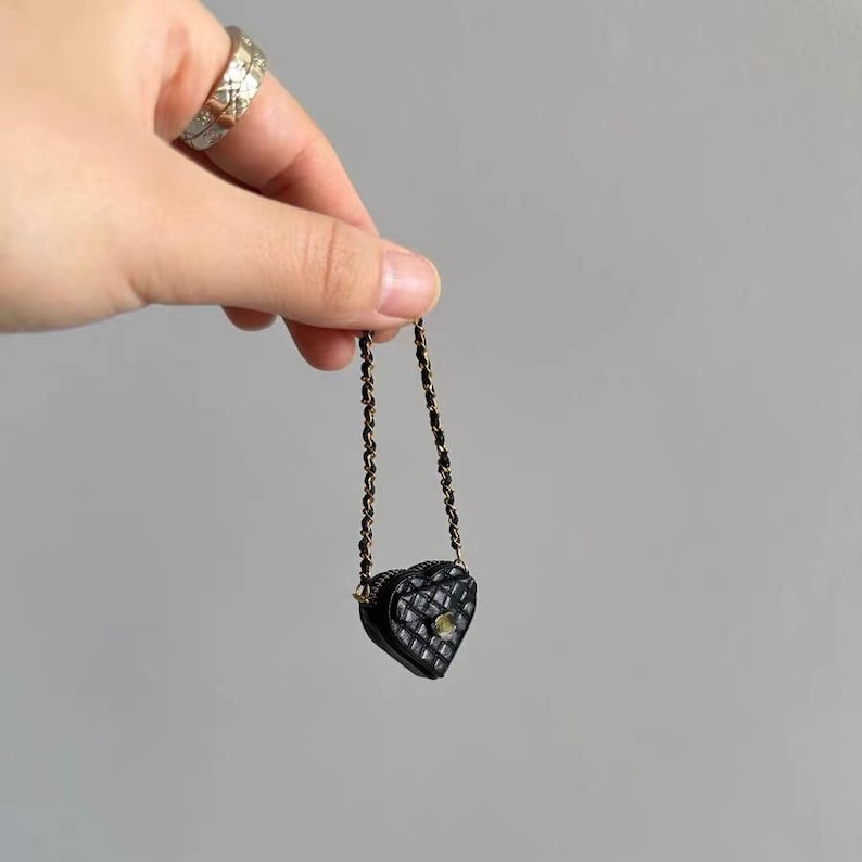 May include: A miniature black heart-shaped purse with a quilted pattern and a gold-colored clasp. The purse has a delicate chain strap. The purse is held up by a hand.