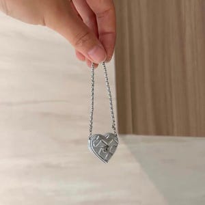 May include: A silver heart-shaped pendant with a chain. The pendant has a textured surface and a small keyhole detail. The chain is delicate and appears to be made of small, linked metal pieces. The pendant is held up by a hand.