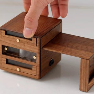 Puede incluir: Un escritorio de madera en miniatura con una superficie de escritorio extraíble y dos cajones. El escritorio es de madera marrón oscuro con perillas de color latón. Una mano está abriendo el escritorio.