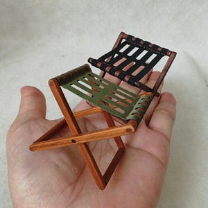 Puede incluir: Silla plegable de madera en miniatura con estructura marrón oscuro y asientos de tela negra y verde oliva. La silla es un modelo a pequeña escala, probablemente para una casa de muñecas. La silla se sostiene en una mano.