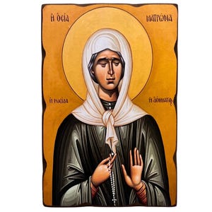 May include: A religious icon featuring a woman with a white head covering and a black robe. The woman is holding a rosary and has a gold halo around her head. The background is a warm, golden color, and the image has text in Greek.