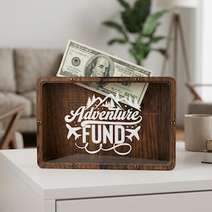 Op de afbeelding: Een rechthoekige houten schaduwdoos met een doorzichtig voorpaneel met de witte tekst Adventure FUND en grafische iconen van vliegtuigen en bergen. Een biljet van honderd dollar is in de bovenste gleuf van de houten houder gestoken.