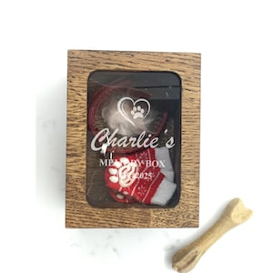 Könnte beinhalten: Eine Holz-Erinnerungsbox mit einer transparenten Frontplatte, die ein rotes Halsband, einen Büschel weißes Fell und eine rot-weiß gestreifte Socke enthält. Die Box ist mit "Charlie's Memory Box" beschriftet. Daneben liegt ein Hundeknochen.