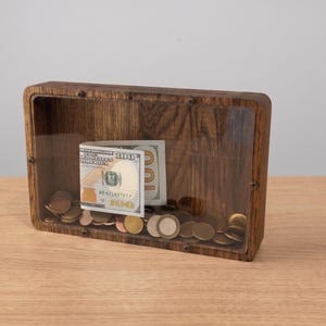 Puede incluir: Una hucha rectangular de madera con un panel frontal de acr&iacute;lico transparente asegurado por cuatro tornillos peque&ntilde;os. Dentro de la caja, hay un billete de cien d&oacute;lares doblado y una colecci&oacute;n de varias monedas met&aacute;licas en el fondo.