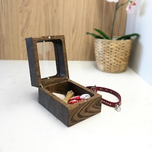 May include: A wooden pet keepsake box with a glass lid, open to show small items. A red leather collar with silver fittings is nearby. The box and collar suggest a pet-themed product.