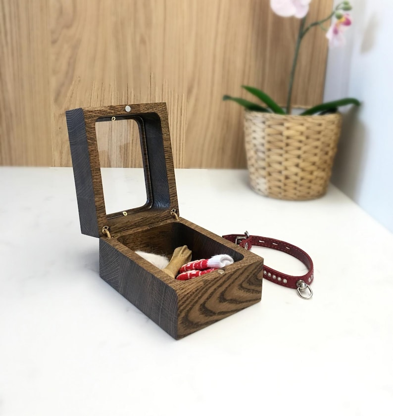 May include: A wooden pet keepsake box with a glass lid, open to show small items. A red leather collar with silver fittings is nearby. The box and collar suggest a pet-themed product.