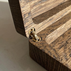 May include: A dark brown wooden box with a visible wood grain texture, featuring an open lid connected by a polished gold-colored metal hinge. The hinge is circular and secured with small screws, showing the interior construction of the box.