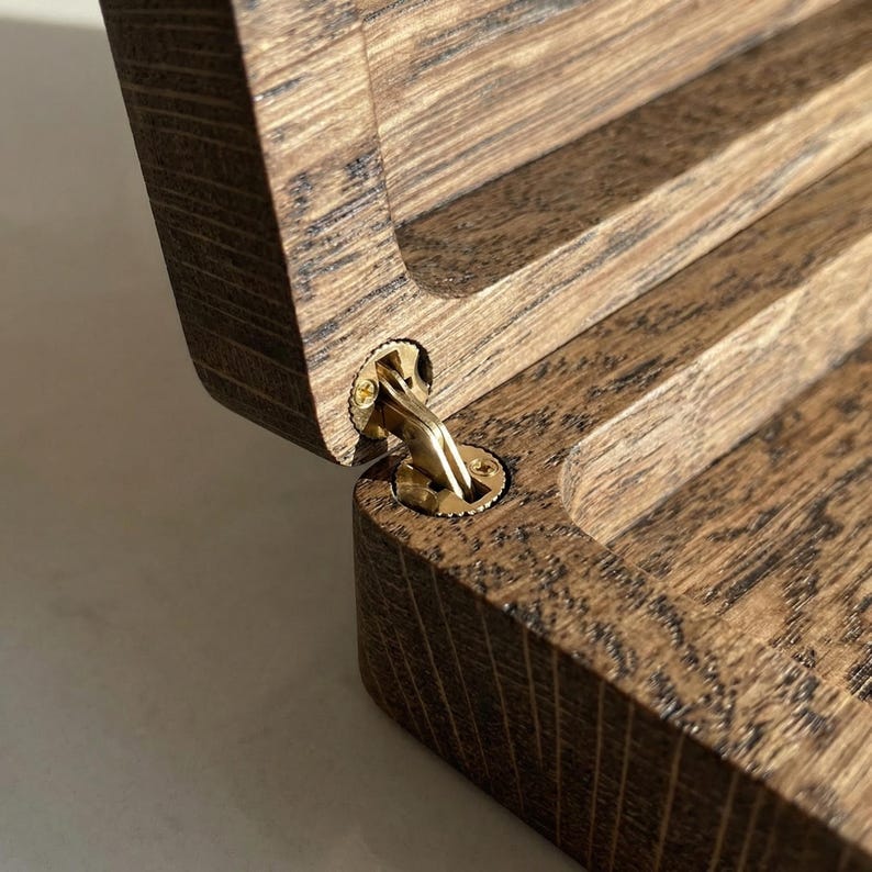 May include: A dark brown wooden box with a visible wood grain texture, featuring an open lid connected by a polished gold-colored metal hinge. The hinge is circular and secured with small screws, showing the interior construction of the box.