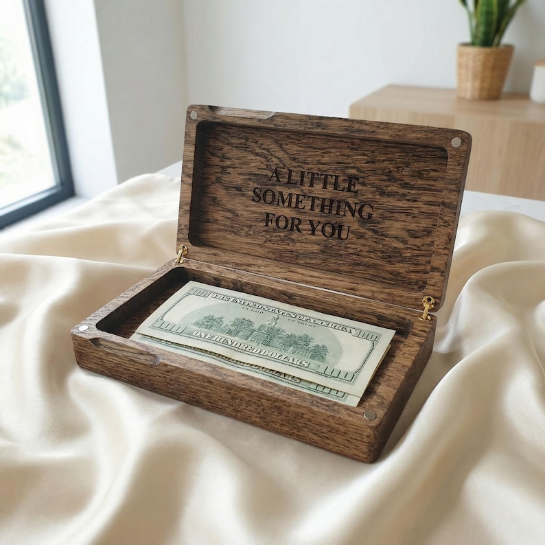 May include: A rectangular dark brown wooden box with a hinged lid. The interior of the lid features the engraved text A LITTLE SOMETHING FOR YOU. Inside the box lies a single one hundred dollar bill. The box has a visible wood grain texture.