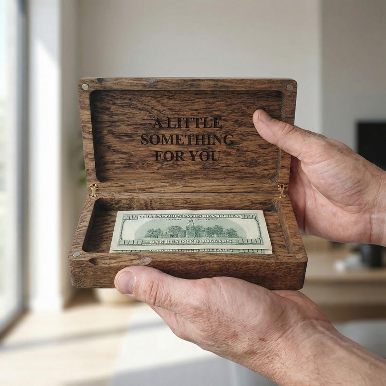 May include: A rectangular dark brown wooden box with a hinged lid. The interior of the lid is engraved with the text A LITTLE SOMETHING FOR YOU. Inside the box lies a single one hundred dollar bill, showing the back side of the currency.