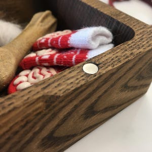 May include: A wooden pet keepsake box containing a bone-shaped chew toy, a small white feather, and a pair of red and white patterned socks. A red collar is visible in the background.