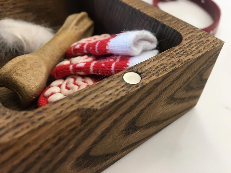 May include: A wooden pet keepsake box containing a bone-shaped chew toy, a small white feather, and a pair of red and white patterned socks. A red collar is visible in the background.