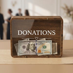 May include: A rectangular wooden donation box with a clear front panel, featuring the white text DONATIONS above a small heart icon and the phrase Thank you for your support. Inside the box are several United States one hundred dollar bills.