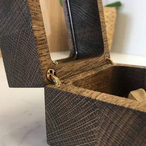 May include: Open wooden box with a hinged lid. The box is made of dark brown wood with a textured grain pattern. The lid is partially open, revealing the interior. The hinges are gold-coloured.
