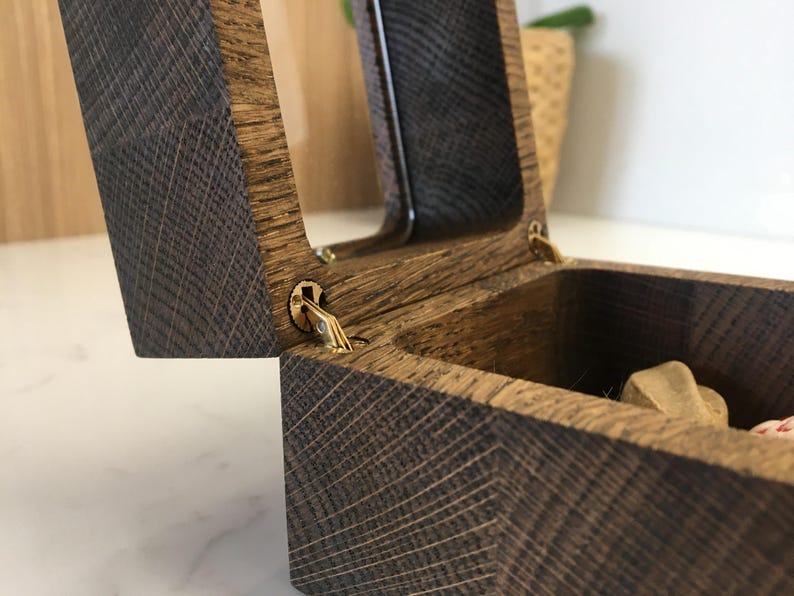 May include: Open wooden box with a hinged lid. The box is made of dark brown wood with a textured grain pattern. The lid is partially open, revealing the interior. The hinges are gold-coloured.