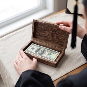 May include: A wooden box with a hinged lid, open to reveal a stack of one hundred dollar bills. The box is being opened by a person wearing a graduation gown. The box is sitting on a neutral colored surface.