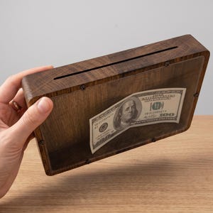 Puede incluir: Una hucha rectangular de madera marr&oacute;n oscuro con un panel frontal de acr&iacute;lico transparente asegurado con peque&ntilde;os tornillos. Una ranura horizontal est&aacute; cortada en el borde superior para insertar dinero, con un billete de cien d&oacute;lares visible.