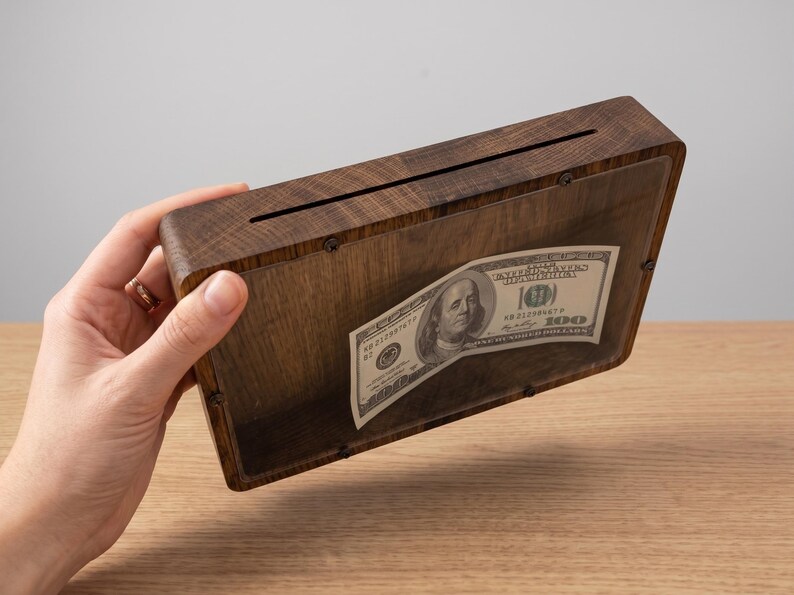 Puede incluir: Una hucha rectangular de madera marr&oacute;n oscuro con un panel frontal de acr&iacute;lico transparente asegurado con peque&ntilde;os tornillos. Una ranura horizontal est&aacute; cortada en el borde superior para insertar dinero, con un billete de cien d&oacute;lares visible.
