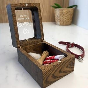 May include: A wooden pet memorial box with a hinged glass lid, containing a small wooden bone, a tuft of white fur, and red and white striped items. A red leather collar with silver studs sits beside the box. The text "2013-2032 MEMORY BOX" is visible on the glass.