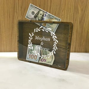 May include: A wooden shadow box with a clear front, containing several U.S. dollar bills. The box is engraved with the names "Meghan & Andrew" inside a wreath design. The box is sitting on a white surface.