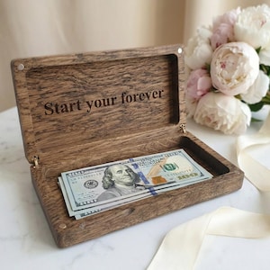 May include: A rectangular dark brown wooden box with a hinged lid, featuring the engraved text "Start your forever" on the inside. The box contains a stack of United States one hundred dollar bills, showcasing the portrait of Benjamin Franklin.