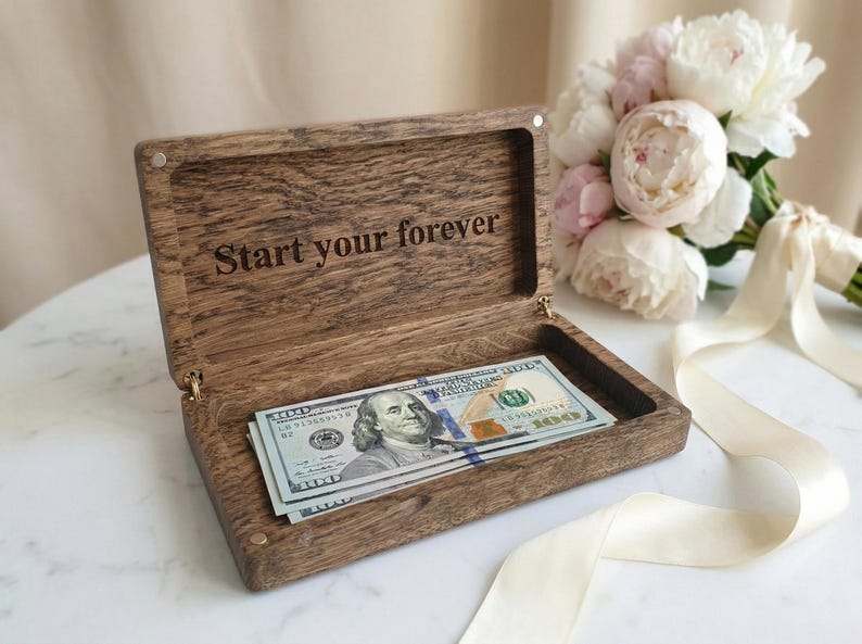 May include: A rectangular dark brown wooden box with a hinged lid, featuring the engraved text "Start your forever" on the inside. The box contains a stack of United States one hundred dollar bills, showcasing the portrait of Benjamin Franklin.
