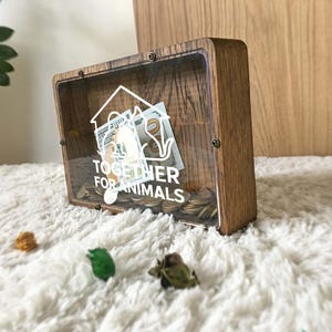 Könnte beinhalten: Eine Holzspardose mit klarer Front, gefüllt mit Münzen und Geldscheinen. Auf der Box steht in weißer Schrift "TOGETHER FOR ANIMALS". Die Box steht auf einer weißen, flauschigen Oberfläche.