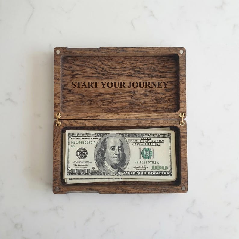 May include: A rectangular dark brown wooden box with a hinged lid, featuring the engraved text START YOUR JOURNEY on the inside of the lid. The box contains a stack of United States one hundred dollar bills neatly placed inside the compartment.
