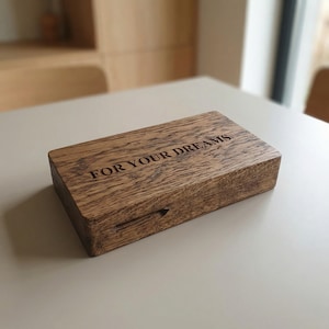 May include: A rectangular wooden box with a dark brown grain pattern. The top surface features the engraved text FOR YOUR DREAMS in a clean, sans-serif font. A small horizontal slot is visible on the front side of the box, near the bottom edge.