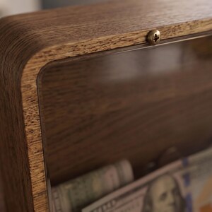 Puede incluir: Una hucha rectangular de madera con un panel frontal de acr&iacute;lico transparente asegurado por un peque&ntilde;o tornillo de metal en la parte superior. El marco presenta un patr&oacute;n de veta de madera visible y el interior contiene varios billetes.
