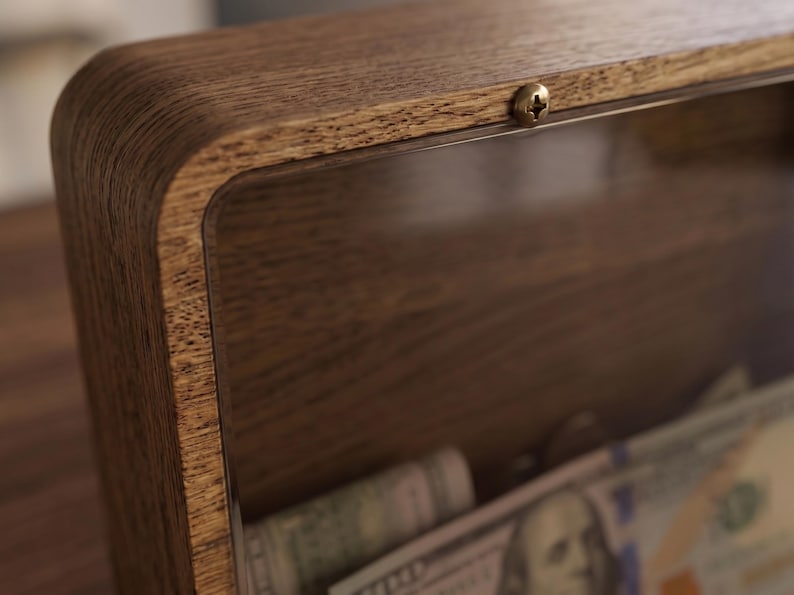 Puede incluir: Una hucha rectangular de madera con un panel frontal de acr&iacute;lico transparente asegurado por un peque&ntilde;o tornillo de metal en la parte superior. El marco presenta un patr&oacute;n de veta de madera visible y el interior contiene varios billetes.