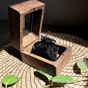 May include: A wooden box with a hinged lid, open to reveal a black velvet pouch inside. The box is made of a dark-stained wood with a rectangular opening. The box is sitting on a woven basket with green leaves.