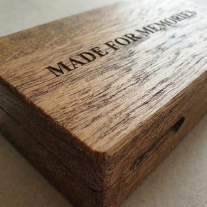 May include: A rectangular wooden box with a natural brown grain finish. The lid features the text MADE FOR MEMORIES engraved in a clear, sans-serif font. The box has a hinged construction and a visible seam running along the side edge.