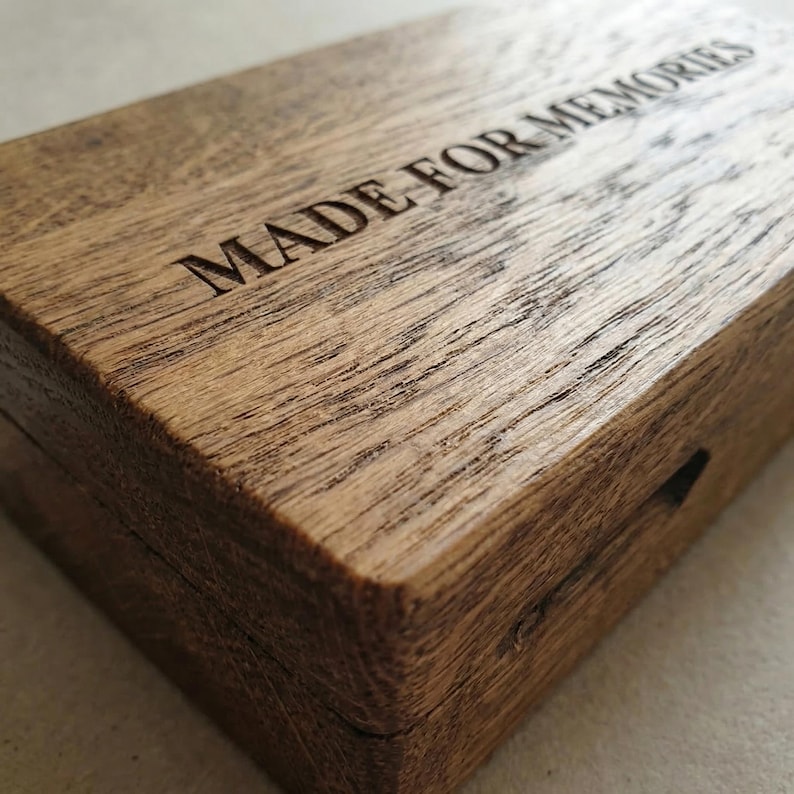 May include: A rectangular wooden box with a natural brown grain finish. The lid features the text MADE FOR MEMORIES engraved in a clear, sans-serif font. The box has a hinged construction and a visible seam running along the side edge.
