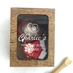 May include: A wooden memory box with a clear front, containing a red collar, a tuft of white fur, and a miniature red and white Christmas stocking. The box is labelled "Charlie's Memory Box 2025". A dog bone is next to the box.