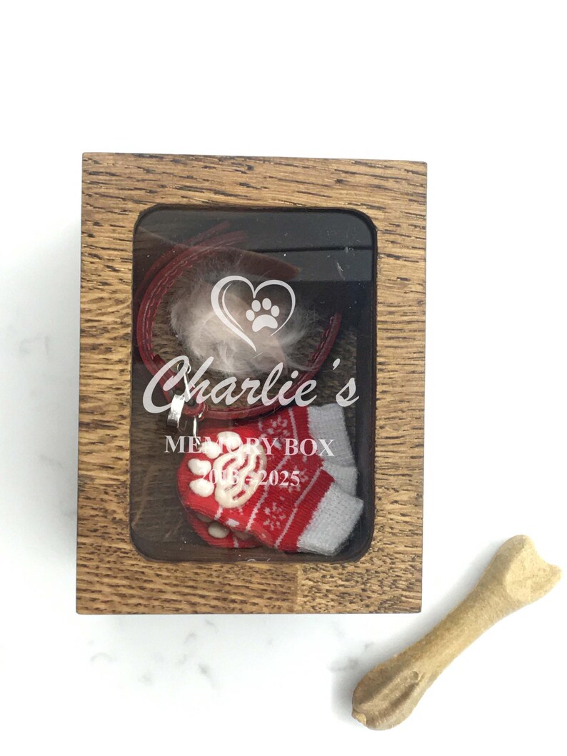 May include: A wooden memory box with a clear front, containing a red collar, a tuft of white fur, and a miniature red and white Christmas stocking. The box is labelled "Charlie's Memory Box 2025". A dog bone is next to the box.