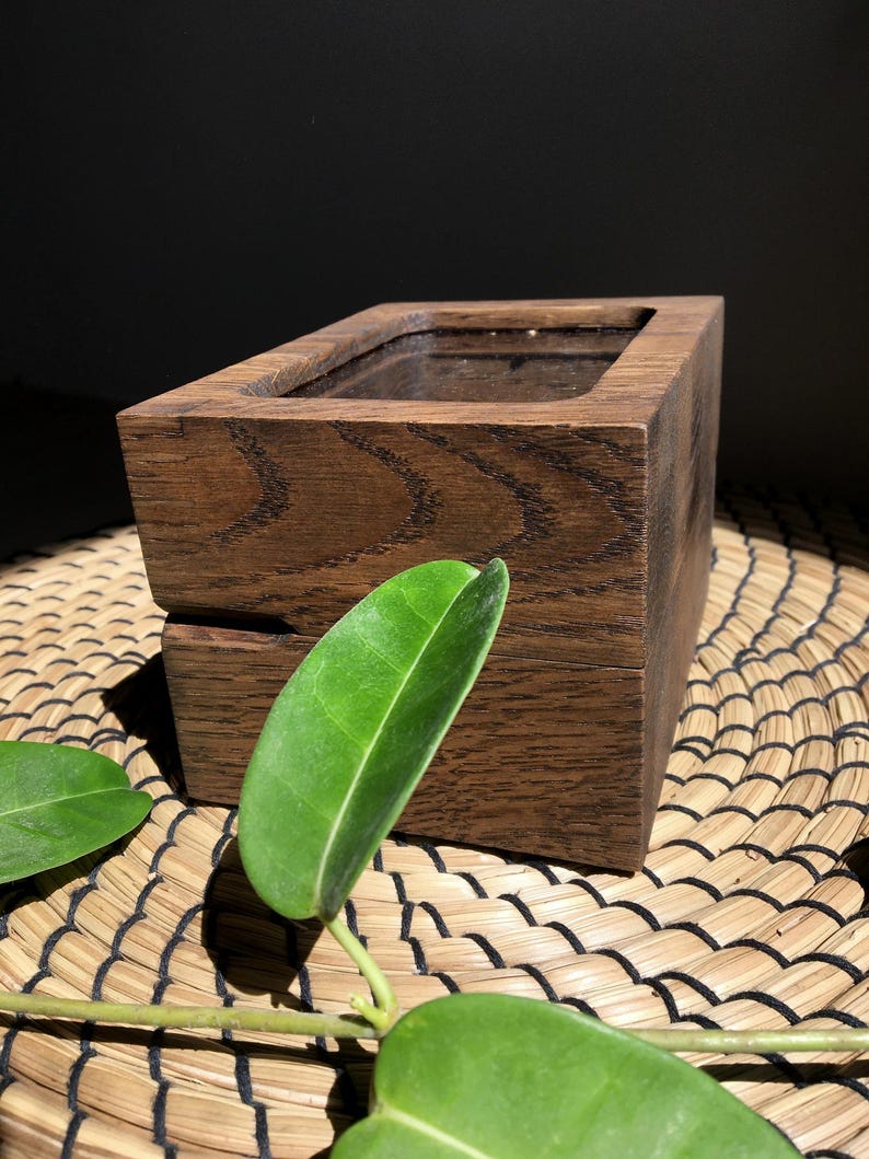 May include: A dark brown wooden box with a rectangular aperture. The box displays a visible wood grain pattern. A green leaf is in the foreground, adding a natural element. The box is likely for storage or display, possibly a decorative item.