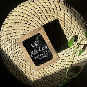 May include: A wooden memory box with a black surface and the inscription "Charlie's MEMORY BOX 2013-2025" in white script. A heart and paw print design is above the name. The box is set on a woven, circular mat.