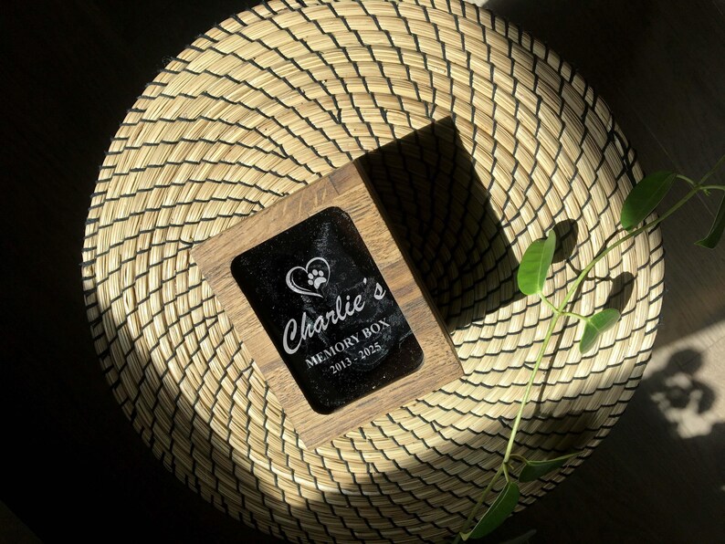 May include: A wooden memory box with a black surface and the inscription "Charlie's MEMORY BOX 2013-2025" in white script. A heart and paw print design is above the name. The box is set on a woven, circular mat.