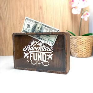 May include: A wooden money bank with a clear front panel, featuring the words "Adventure FUND" in white script, with a mountain and airplane graphic. A one hundred dollar bill is partially visible inside the bank.