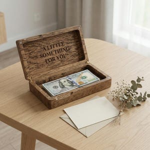 May include: A rectangular wooden box with a hinged lid, featuring the engraved text A LITTLE SOMETHING FOR YOU on the inside. The box contains a stack of United States one hundred dollar bills, and a blank cream-colored envelope sits nearby.