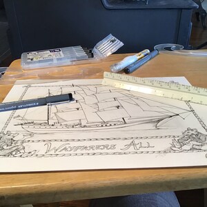 May include: A black and white drawing of a ship with the text "Wayfarers All" on a white sheet of paper. The drawing is surrounded by a rope border. A ruler and a black pen are on the table next to the drawing.