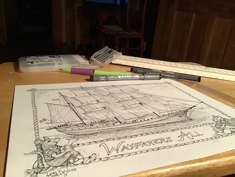 May include: A black and white ink drawing of a ship with the text "Wayfarers All" on a white sheet of paper. The drawing is surrounded by a rope border. There are various art supplies on the table, including a ruler, a purple pen, and black markers.